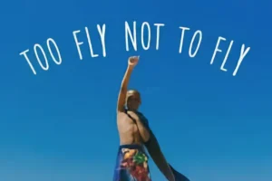 Person standing outdoors with one arm raised, wrapped in a colorful towel, under a clear blue sky with the text "TOO FLY NOT TO FLY" above them.