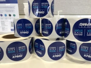 Several rolls of blue KPMG stickers with the text "It starts with a book and ends with a story" are stacked on a table.