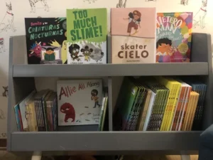 A gray bookshelf holds children's picture books, some in English and some in Spanish, displayed facing forward and arranged upright on two shelves.