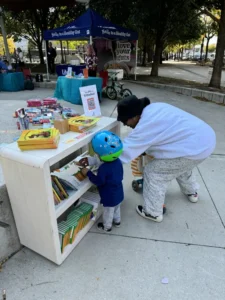 An adult and child wearing a helmet look through books on a white bookshelf outdoors, with informational booths and tables set up in the background.