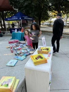 Two girls look at colorful children's books displayed on tables outdoors; a man stands nearby and informational booths are set up in the background.