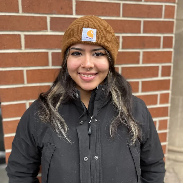 A person wearing a brown beanie and black jacket stands in front of a red brick wall, smiling at the camera.