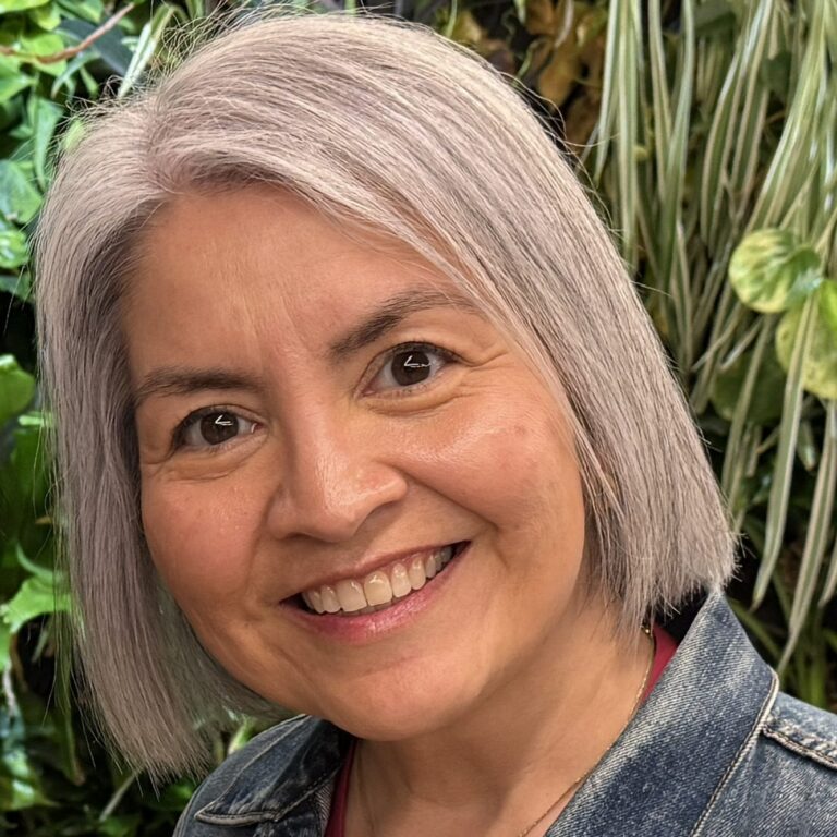 A smiling woman with gray hair and a denim jacket stands in front of a wall of green plants.