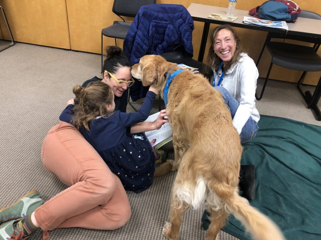 Two women and a young girl are sitting on the floor, smiling and interacting with a large, golden dog in an indoor setting.