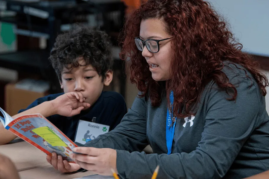 An adult with curly hair reads "If I Ran the Zoo" by Dr. Seuss to a young boy in a classroom. The boy listens attentively, resting his chin on his hand.