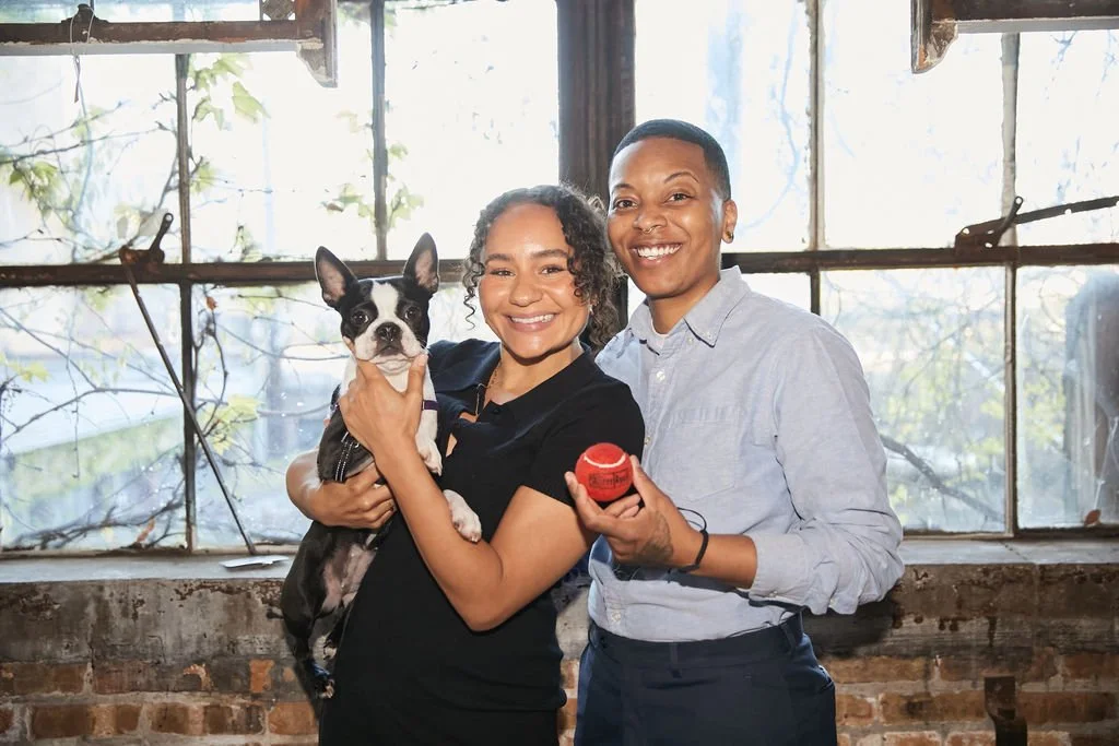 Two people stand indoors by a window; one holds a small black and white dog, and the other holds a red ball.