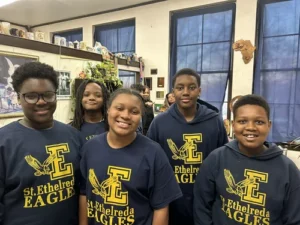 Five children wearing matching "St. Ethelreda Eagles" shirts stand together and smile in a classroom decorated with art and various items.