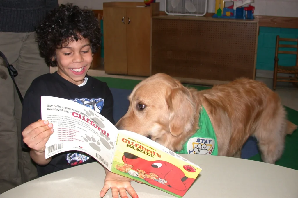 A smiling child sits at a table holding a Clifford book, while a golden retriever wearing a green vest looks at the pages.