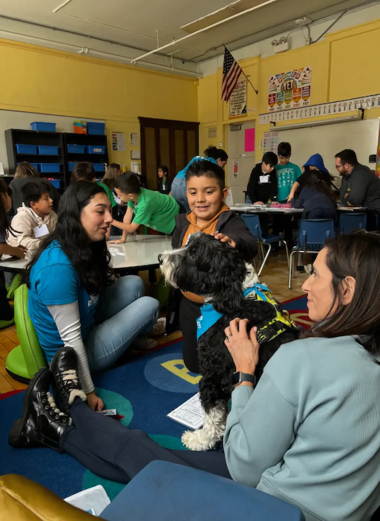 Children and adults interact with a therapy dog in a classroom setting, sitting on the floor and at tables, with educational materials visible in the background.
