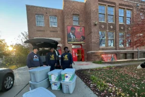 Three people stand beside a car with its trunk open, unloading plastic storage bins in front of a brick school building with a red door.