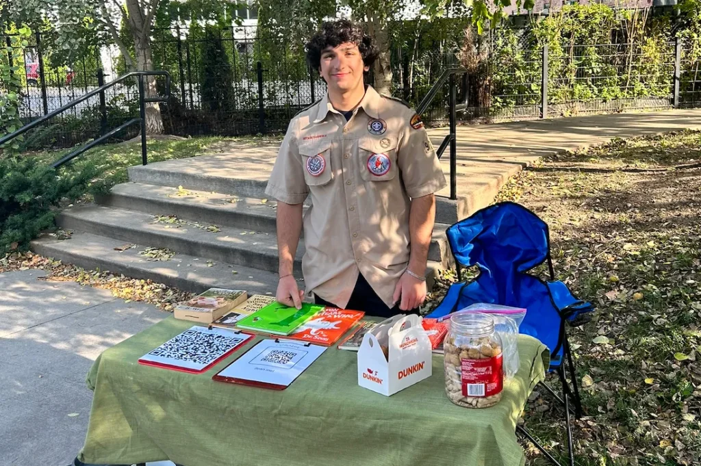 A person in a scout uniform stands behind a table with books, papers, a QR code, a jar of cookies, and Dunkin’ boxes, set up outdoors near a staircase and trees.