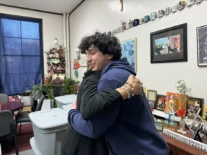 Two people are hugging in a room decorated with framed photos, mugs, plants, and various personal items.