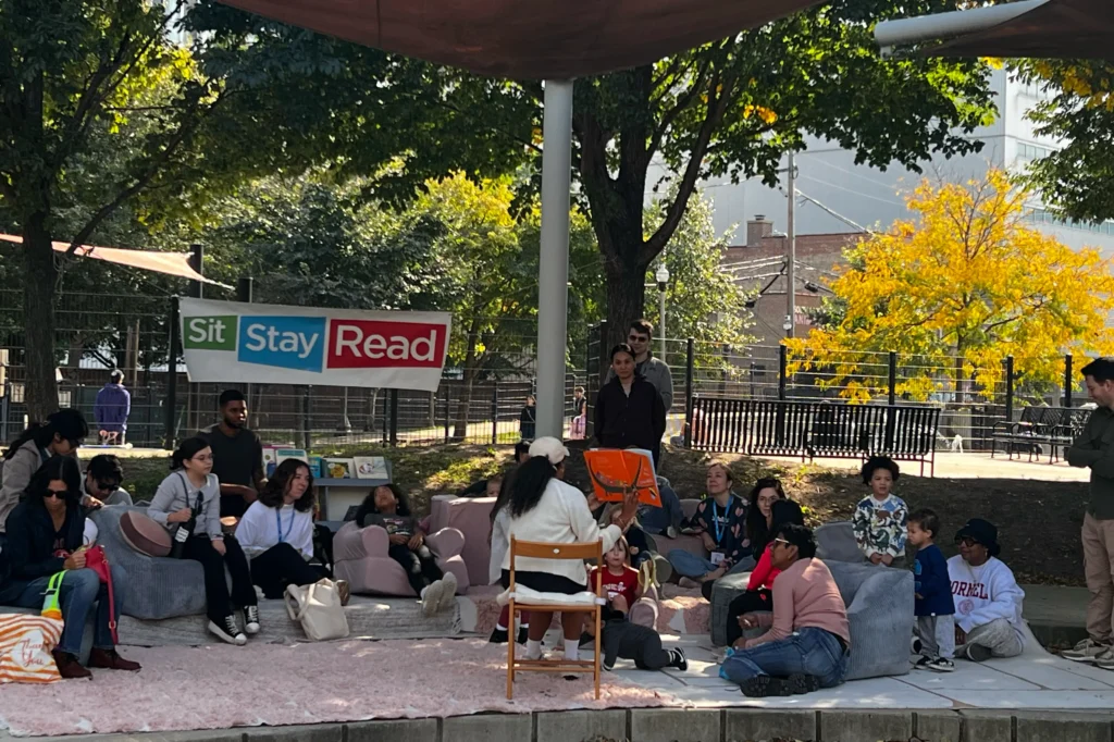 A group of adults and children sit outdoors on cushions and chairs, listening to a person reading aloud under a "Sit Stay Read" banner in a shaded area with trees.