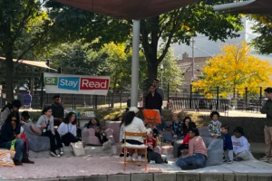 A group of adults and children sit outdoors on cushions and chairs, listening to a person reading aloud under a "Sit Stay Read" banner in a shaded area with trees.