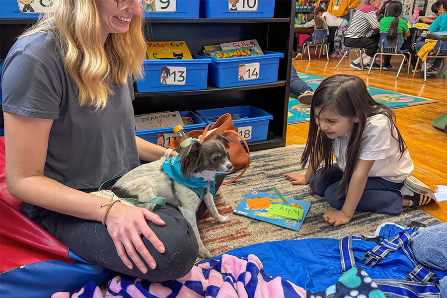A woman and a child sit on the floor next to a small dog wearing a blue bandana, with books and storage bins visible in the background.