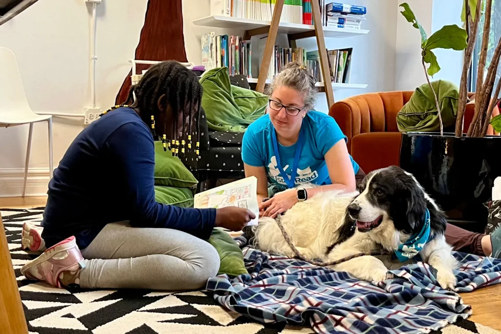 A girl reads a book to an adult woman and a large black-and-white dog lying on a blanket in a cozy room with bookshelves and plants.