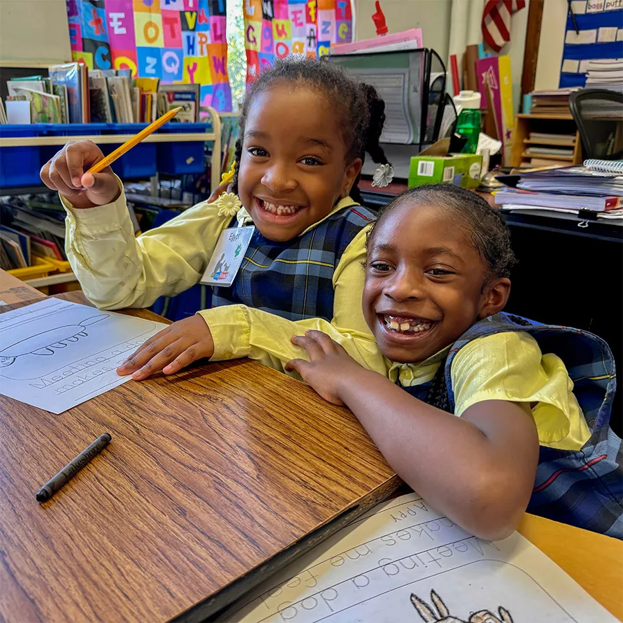 Two young girls in school uniforms smile while sitting at a classroom desk with worksheets, a pencil, and a crayon. Colorful educational posters are visible in the background.