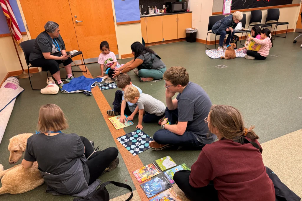Adults and children sit on the floor in a classroom, reading books and interacting with therapy dogs.