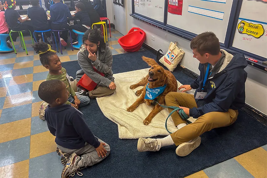 Three children and two adults sit on the floor around a therapy dog in a classroom. The dog is on a blanket wearing a blue bandana. Classroom desks and a whiteboard are in the background.