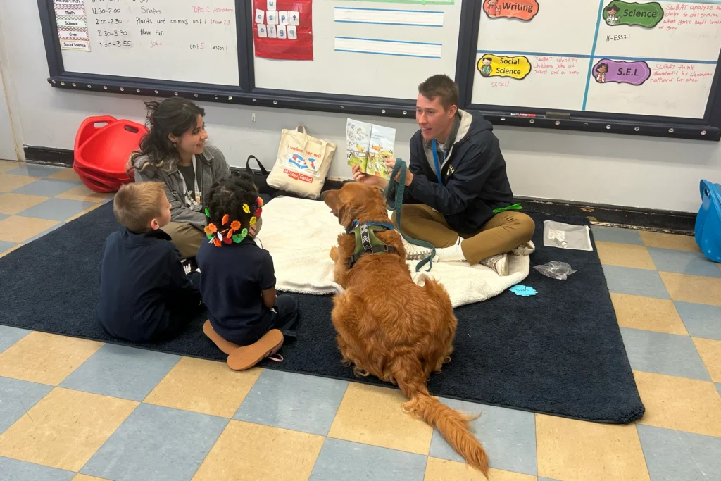 An adult reads a book to two children and another adult while a golden retriever lies on a rug in a classroom. Educational materials are visible on the wall and floor.