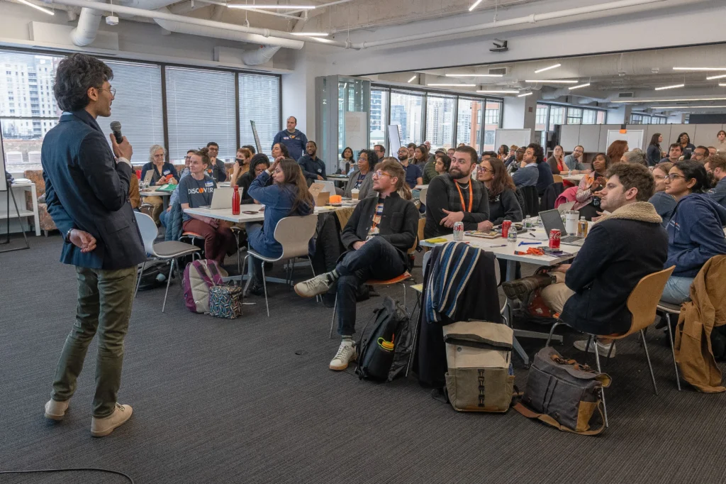 A person stands and speaks to a large group of people seated at tables in a modern, well-lit conference room.