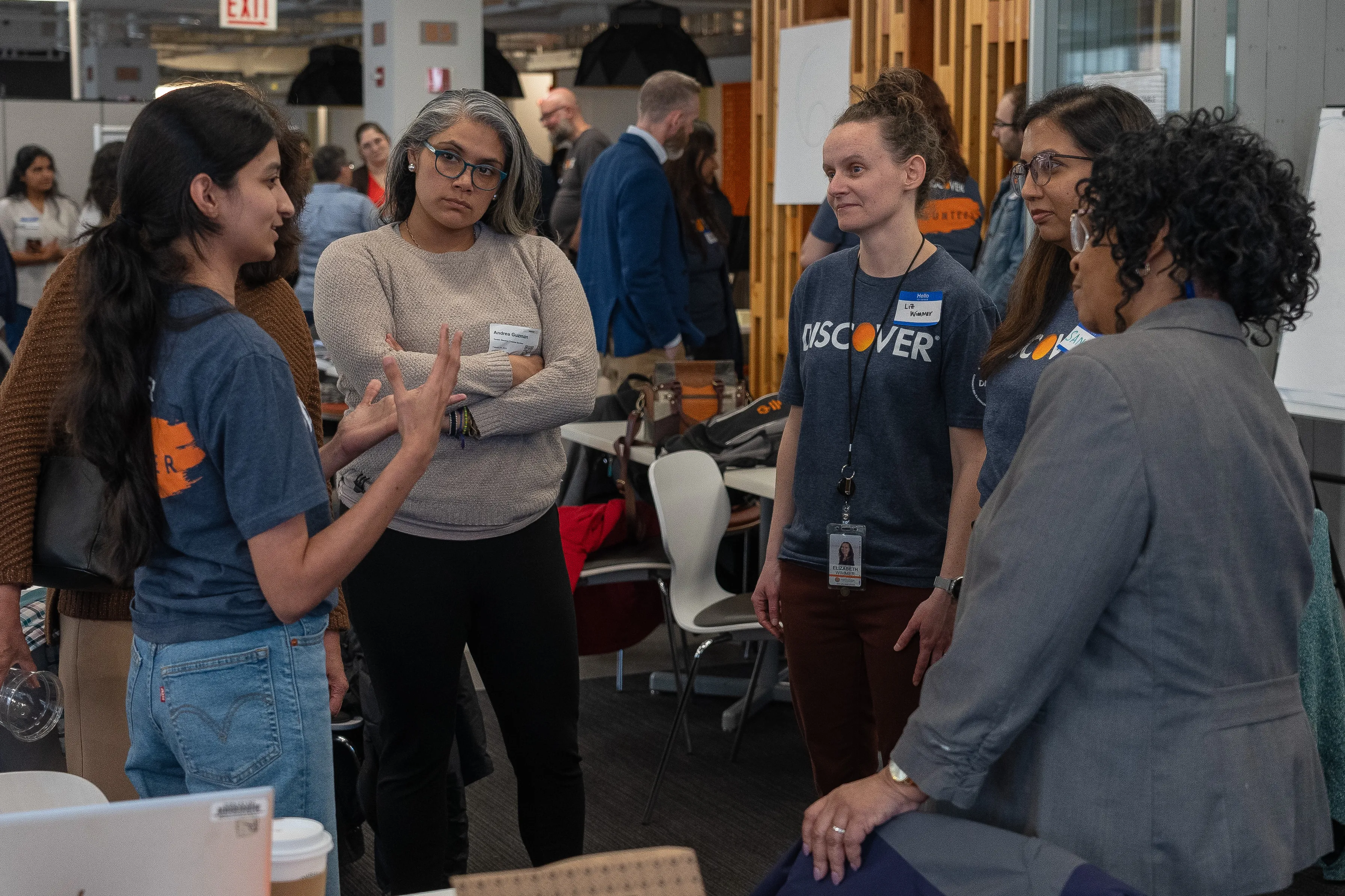 A group of five women, some wearing "Discover" shirts, stand in a circle having a conversation in an office setting with others in the background.