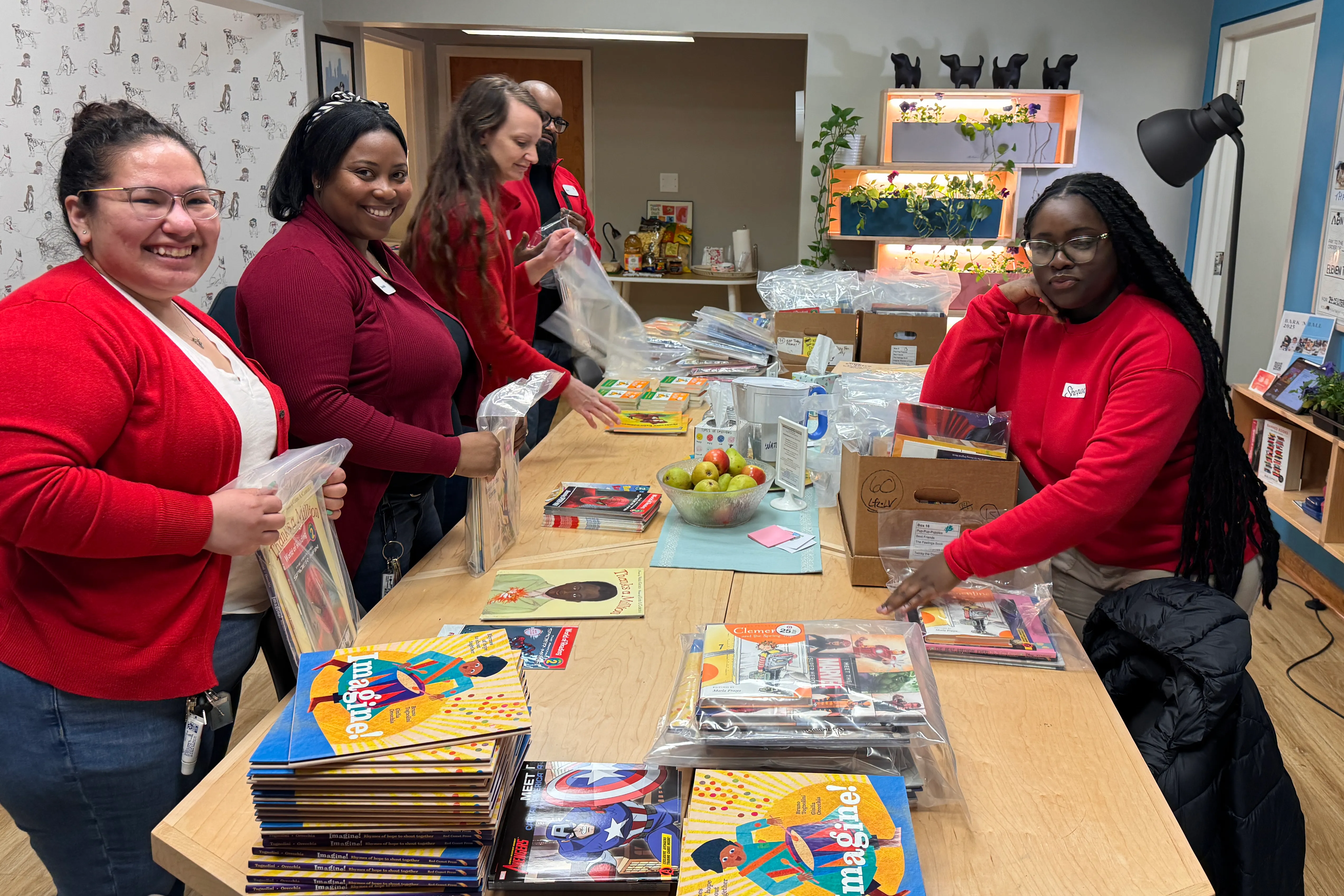 Four women stand around a table sorting and packing colorful children's books into plastic bags in a brightly lit room.
