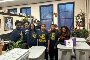 A group of students and an adult pose in a classroom with large storage bins labeled "Thank You Eagle Scout Grant," plants, and computers visible in the background.