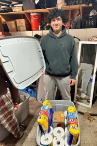A person in a gray hoodie stands in a garage, holding the lid of a storage bin filled with cleaning supplies and paper towels. Shelves and miscellaneous items are in the background.