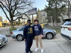 Two young men stand on a sidewalk in front of parked cars and trees, both wearing school sweatshirts. A school sign and building are visible in the background.