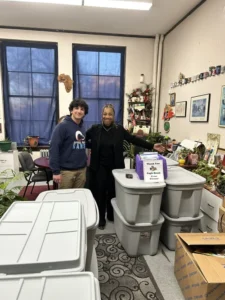 Two people stand in a cluttered room behind a stack of large gray storage bins, with a sign thanking Eagle Scouts attached to one of the bins.