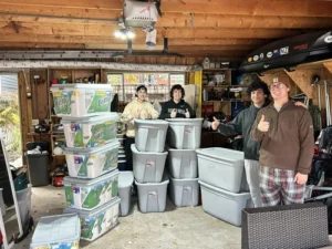 Four people stand in a garage, smiling and giving thumbs up, next to tall stacks of plastic storage bins and boxes. Various items and tools are visible in the background.