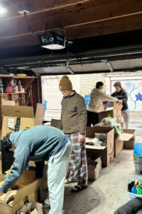 Four people sort and pack items into cardboard boxes in a garage, with household supplies and containers visible around them.