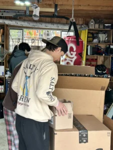 Two people stand in a garage, one wearing a "Salty Crew" hoodie and cap, unpacking large cardboard boxes on a table. The garage is filled with various tools and items.