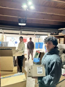 Three people stand in a garage filled with cardboard boxes; two are holding boxes while the third stands in the background near the open garage door.