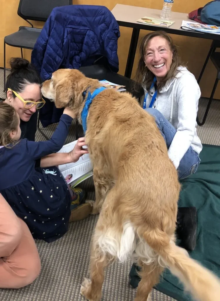 A large dog interacts with two women and a child sitting on the floor in a room; one woman is smiling at the camera.