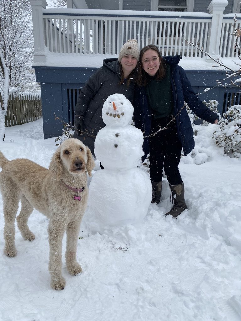 Two people and a dog stand in the snow next to a snowman with a carrot nose, rock eyes, and button mouth in front of a house.