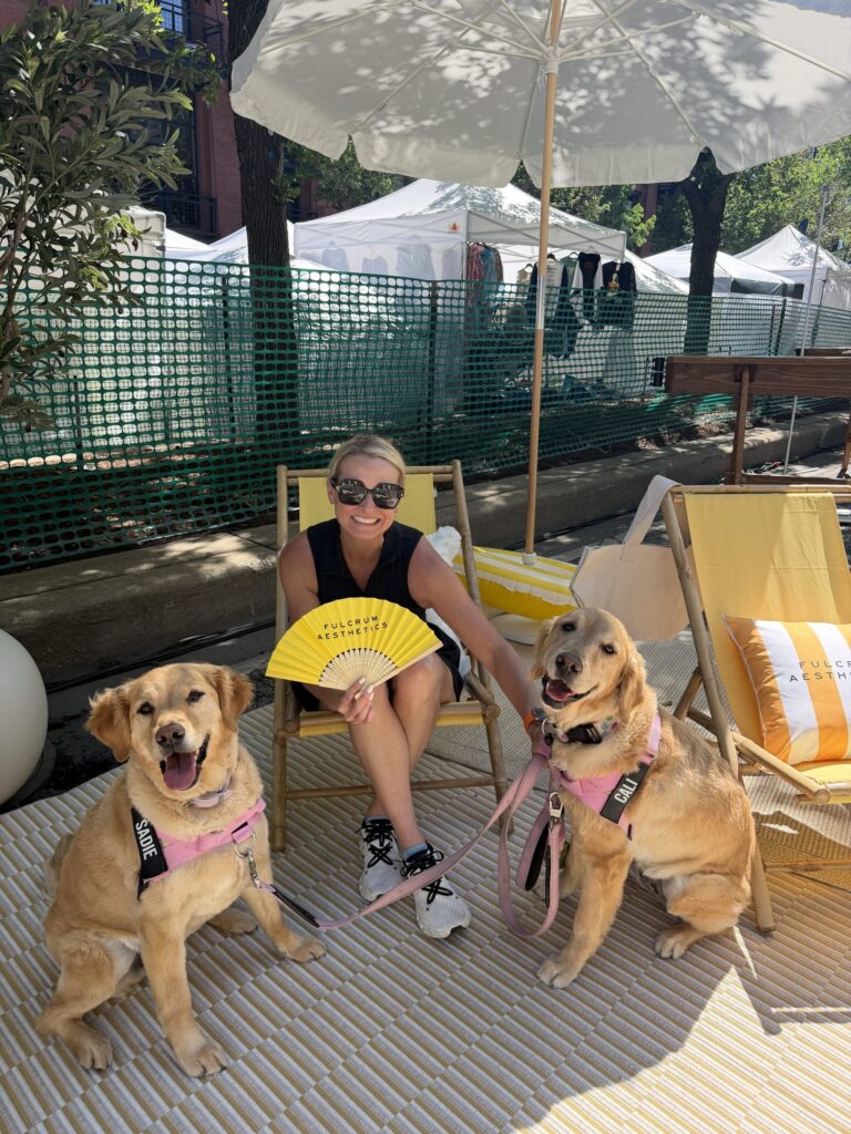 Woman sitting on a lounge chair under an umbrella, holding a yellow fan, with two leashed golden retrievers wearing harnesses, at an outdoor event with tents in the background.
