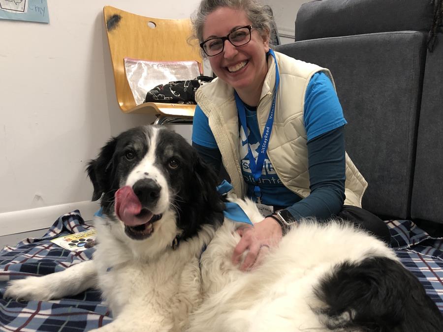 A woman wearing glasses and a blue shirt sits on a plaid blanket, smiling, beside a large black and white dog that is licking its nose.