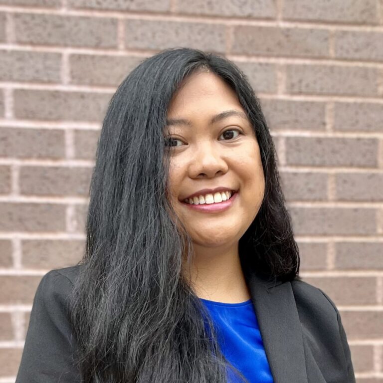 Woman with long black hair, wearing a black blazer over a blue top, stands in front of a brick wall, smiling at the camera.